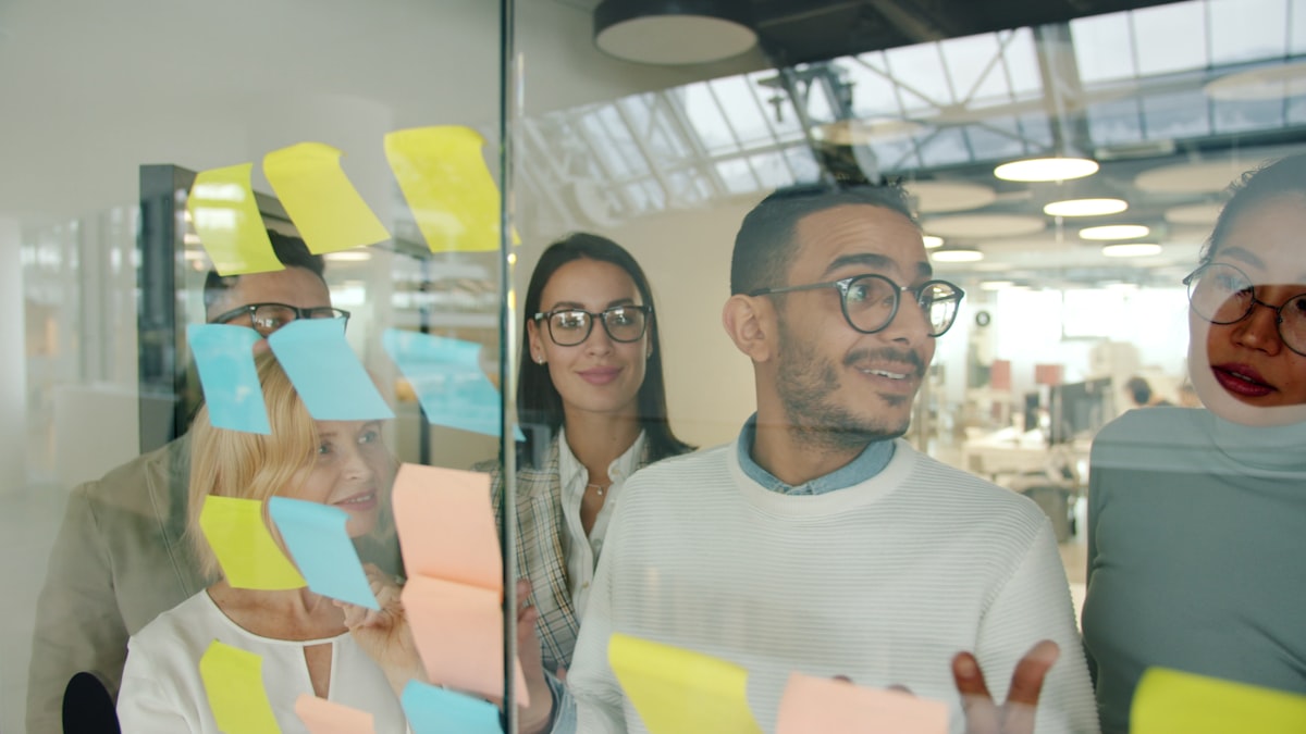 Adultes multiethniques travaillant ensemble, homme animant une séance de brainstorming avec des collègues devant un tableau couvert de post-its ambiance de travail engagée et collaborative